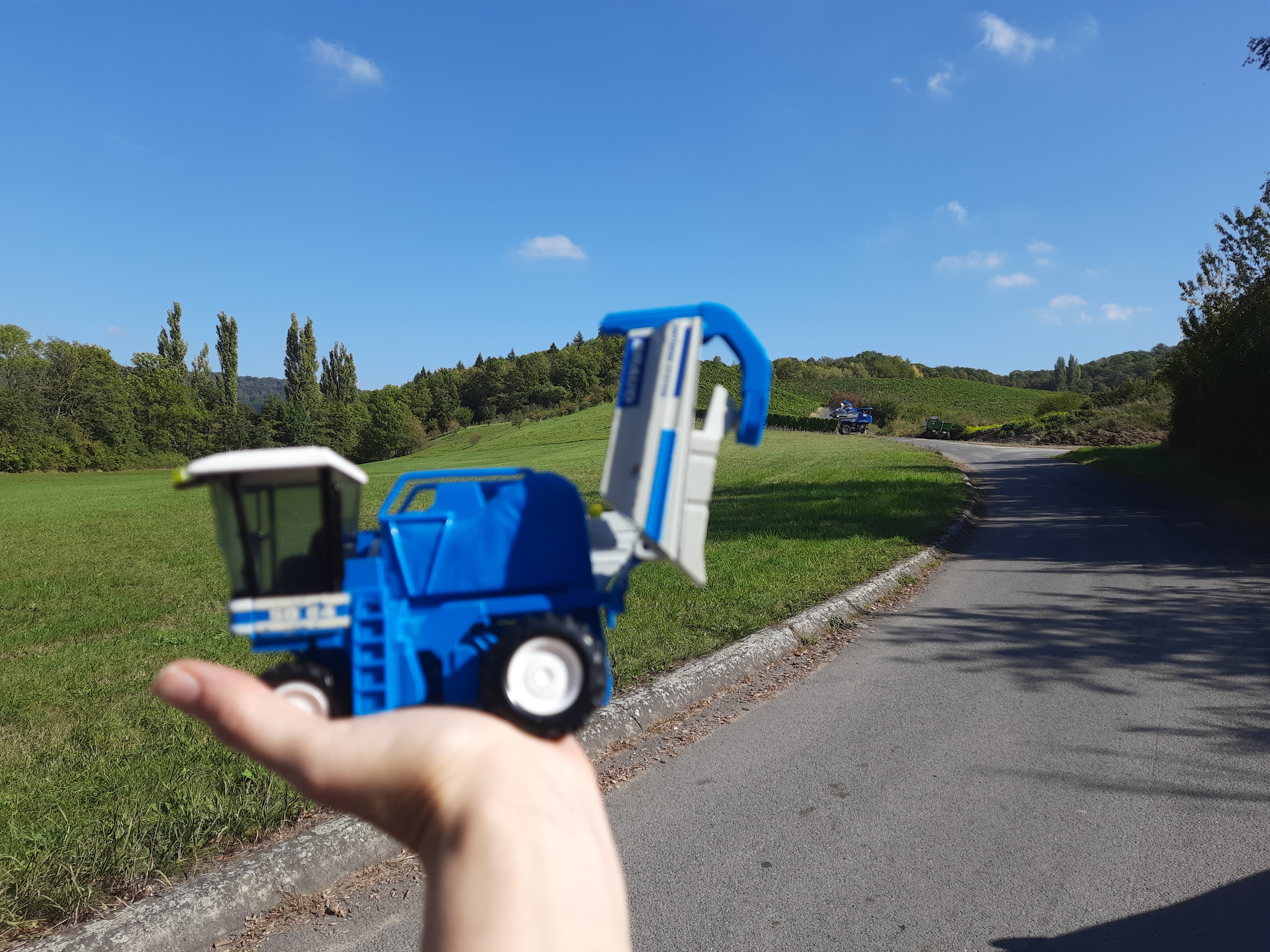 Eine Hand hält einen kleinen blauen Spielzeugbagger vor die Landschaft.